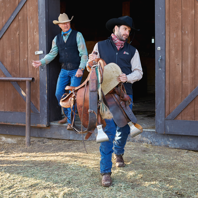 A man wearing a Justin vest carrying a saddle walking out of a barn in the snow.